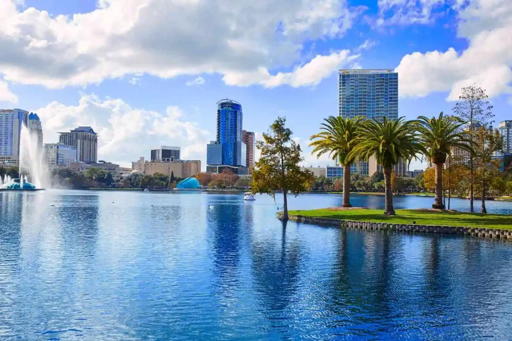 A scenic view of the Orlando skyline with tall buildings reflected on a calm lake. The foreground features palm trees and a small grassy island, while a fountain sprays water high into the air under Florida's partly cloudy blue sky.