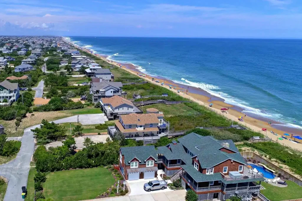 An aerial view of a coastal town in North Carolina's Outer Banks shows houses lining the shore. The beach has a few sunbathers and umbrellas, while waves gently crash onto the sandy shore. Greenery surrounds the residential area, and the ocean stretches to the horizon under a blue sky with few clouds.