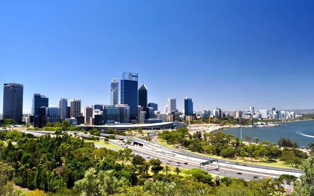 A vibrant cityscape of Perth, Australia, showcases modern high-rise buildings against a clear blue sky. Lush greenery and a multi-lane highway adorn the foreground, while the river gracefully curves along the right. The sunny day accentuates Perth's stunning architecture.