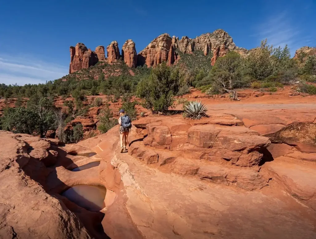 A hiker wearing a backpack walks on the rugged, rocky terrain of Soldier Pass Trail with small pools of water. Tall, red rock formations and lush green vegetation surround the area. A clear blue sky stretches above the scenic landscape.