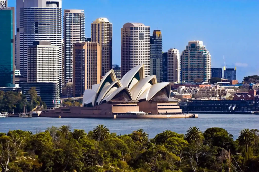 Sydney Opera House with its iconic sail-like design is prominently featured in the foreground, set against a backdrop of Sydney's tall modern skyscrapers. The scene is framed by the clear blue sky above and lush green trees in the foreground.