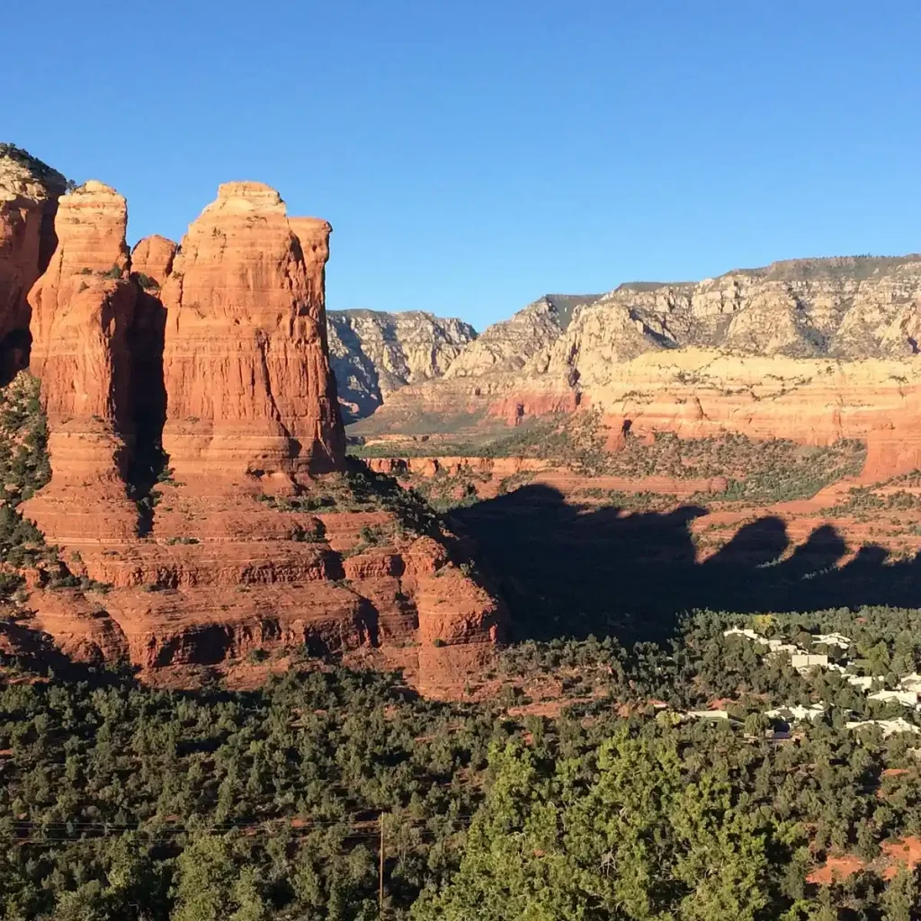 A stunning landscape of Sedona, Arizona, featuring towering red rock formations under a clear blue sky. The foreground shows lush vegetation, while the background reveals rugged, rocky cliffs. A small cluster of buildings is visible at the base of the rocks near the Teacup Trail to Coffeepot Rock.