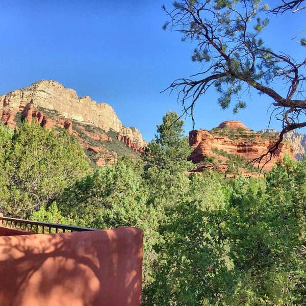 A scenic view of Sedona's red rock formations, topped with lush green vegetation under a clear blue sky, unfolds near the Yavapai Vista Trail. In the foreground, a portion of a red stucco wall is visible, partially shaded by trees. Pine branches frame the image on the right.