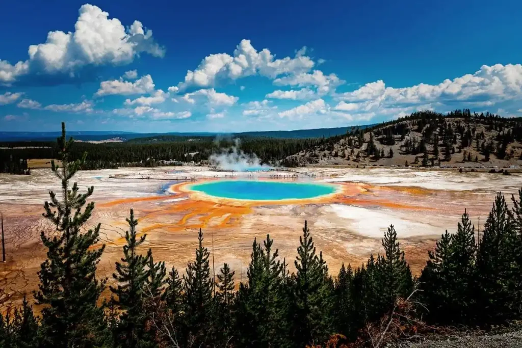 A vibrant, multicolored hot spring in Yellowstone National Park is surrounded by evergreen trees, set against a backdrop of hills and a blue sky with scattered clouds. The hot spring showcases hues of blue, green, yellow, and orange, creating a striking contrast with Wyoming's brown, rocky terrain.