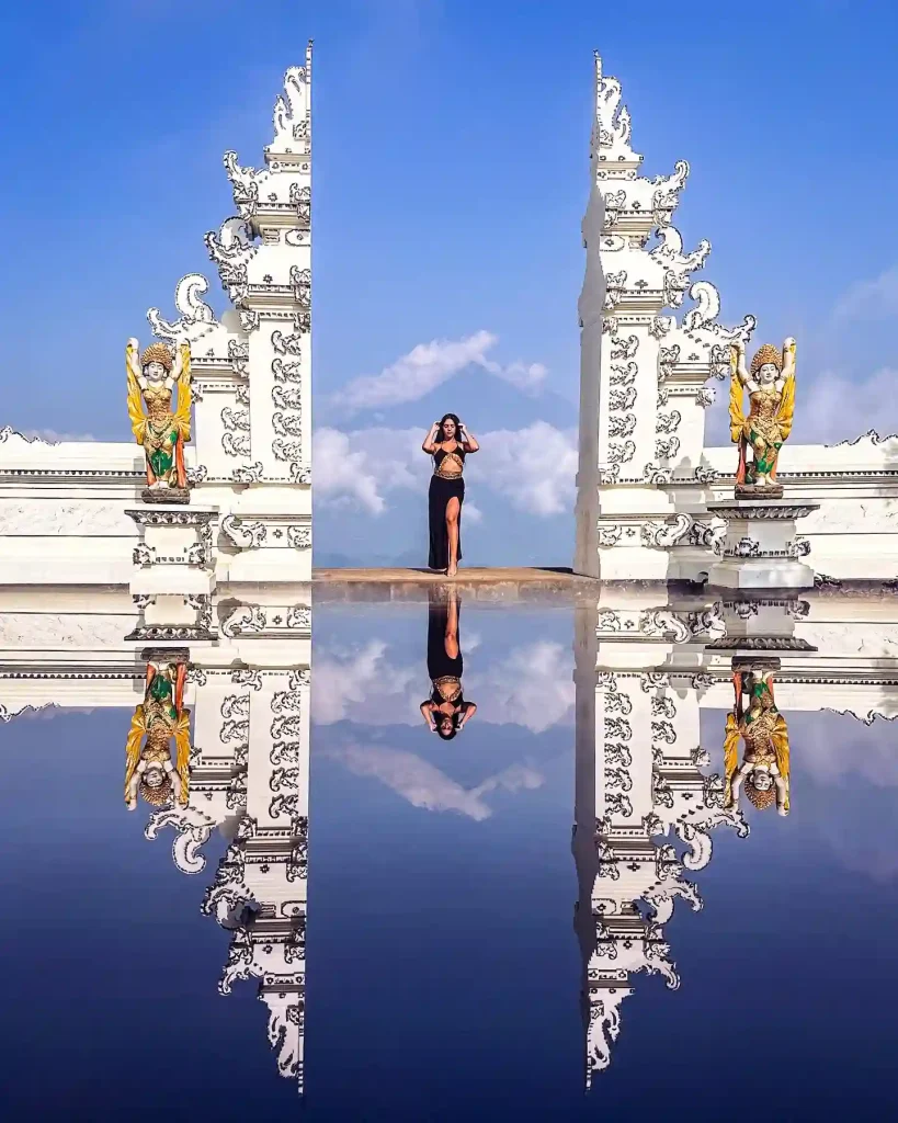 A woman stands between two intricately carved white temple gates in Bali, mirrored by a reflective surface below. She poses against a bright blue sky with clouds, flanked by two vibrant guardian statues. The scene creates a symmetrical, ethereal effect.