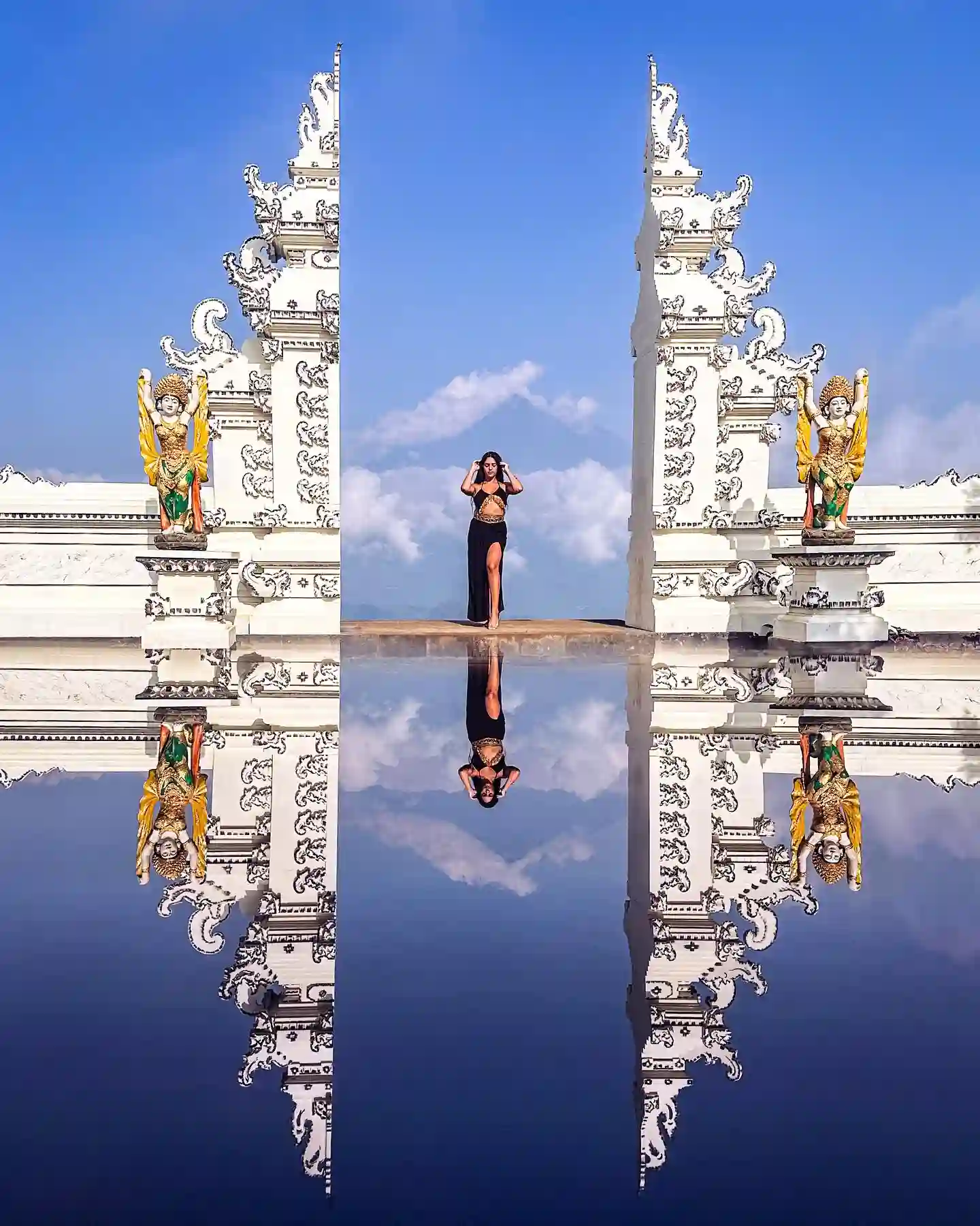 A woman stands between two intricately carved white temple gates in Bali, mirrored by a reflective surface below. She poses against a bright blue sky with clouds, flanked by two vibrant guardian statues. The scene creates a symmetrical, ethereal effect.