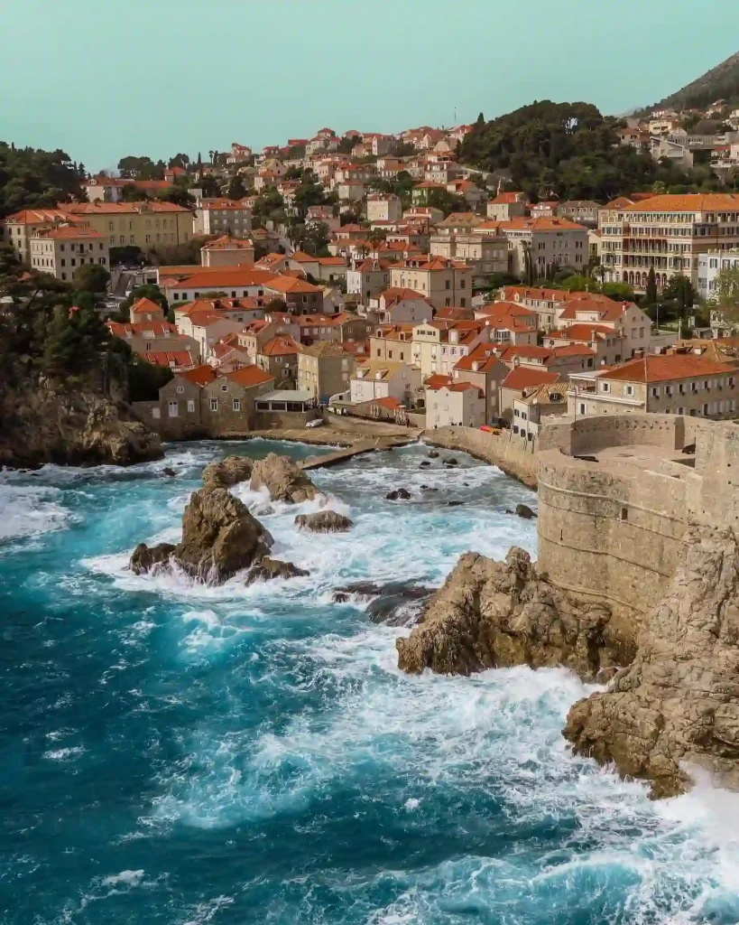 A coastal town with numerous orange-roofed buildings is seen against a backdrop of green hills. Situated along a rocky coastline in Croatia, with waves crashing against the shore, Dubrovnik's stone fortification extends into the water, adding to the picturesque scenery.