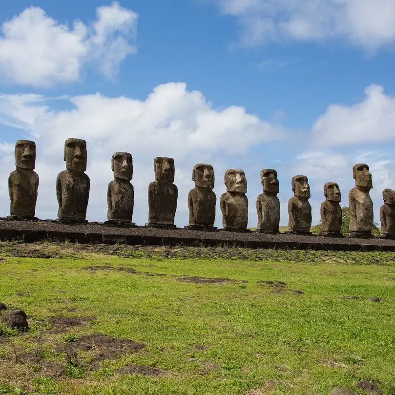 A line of Moai statues stands on a stone platform under a partly cloudy sky on Easter Island, Chile. The statues, carved from volcanic tuff, face inland, displaying stoic expressions. The foreground features grassy terrain with scattered rocks.