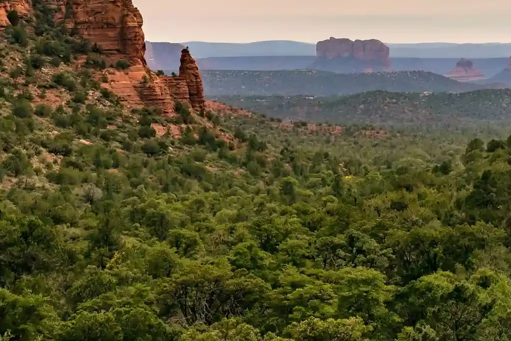 A scenic view of a lush green valley surrounded by rocky red cliffs, with a distant range of mountains under a cloudy sky. The landscape along the Fay Canyon Trail has a combination of greenery and arid rock formations, creating a serene and picturesque setting.