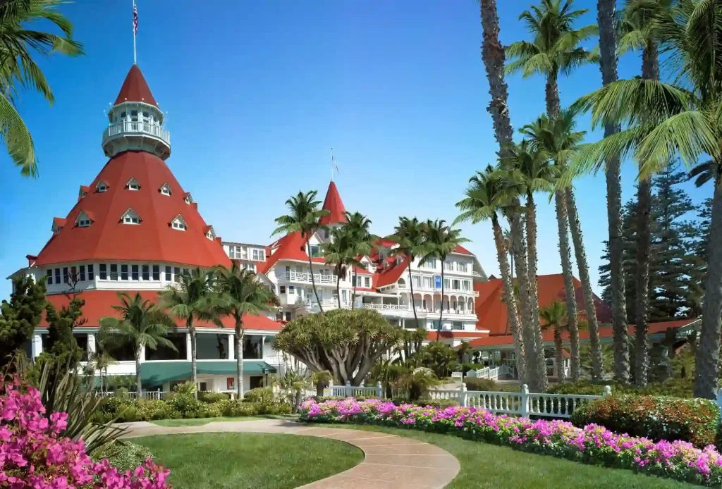 The iconic Hotel del Coronado, with its historic red roofs and white trim, is surrounded by palm trees, vibrant flowers, and a lush lawn under a clear blue sky.