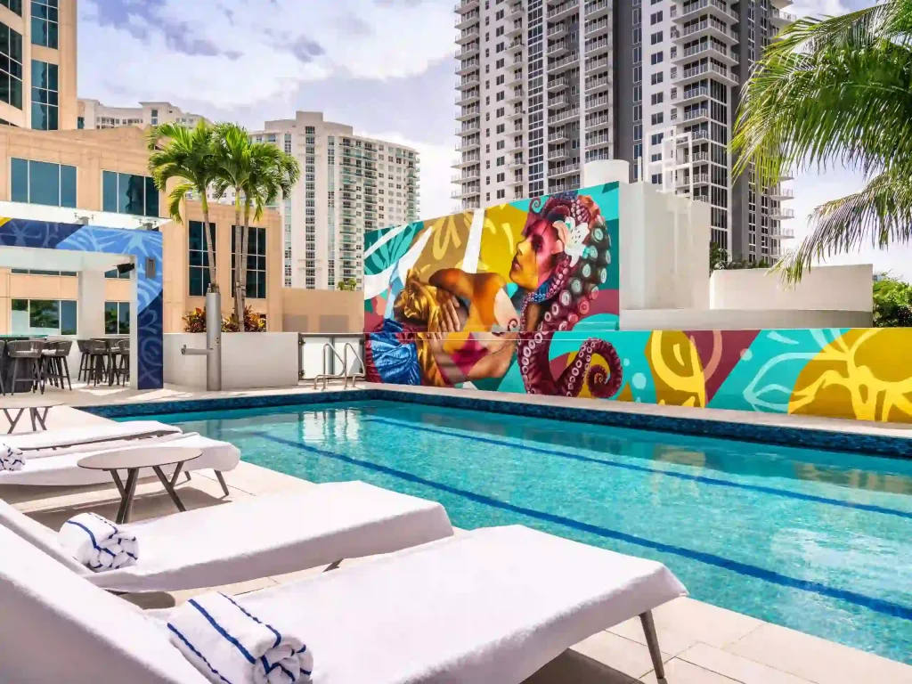 The outdoor swimming pool area at Hyatt Centric Las Olas Fort Lauderdale boasts several lounge chairs and a vibrant mural depicting abstract art and human figures, surrounded by tall modern buildings.