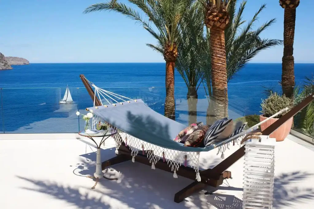 A hammock with decorative pillows is set up on a white patio overlooking the clear blue ocean of Ibiza. Palm trees provide shade, and a sailboat is visible in the distance on the water. A glass railing borders the patio, offering an unobstructed view of this serene seascape in Spain.