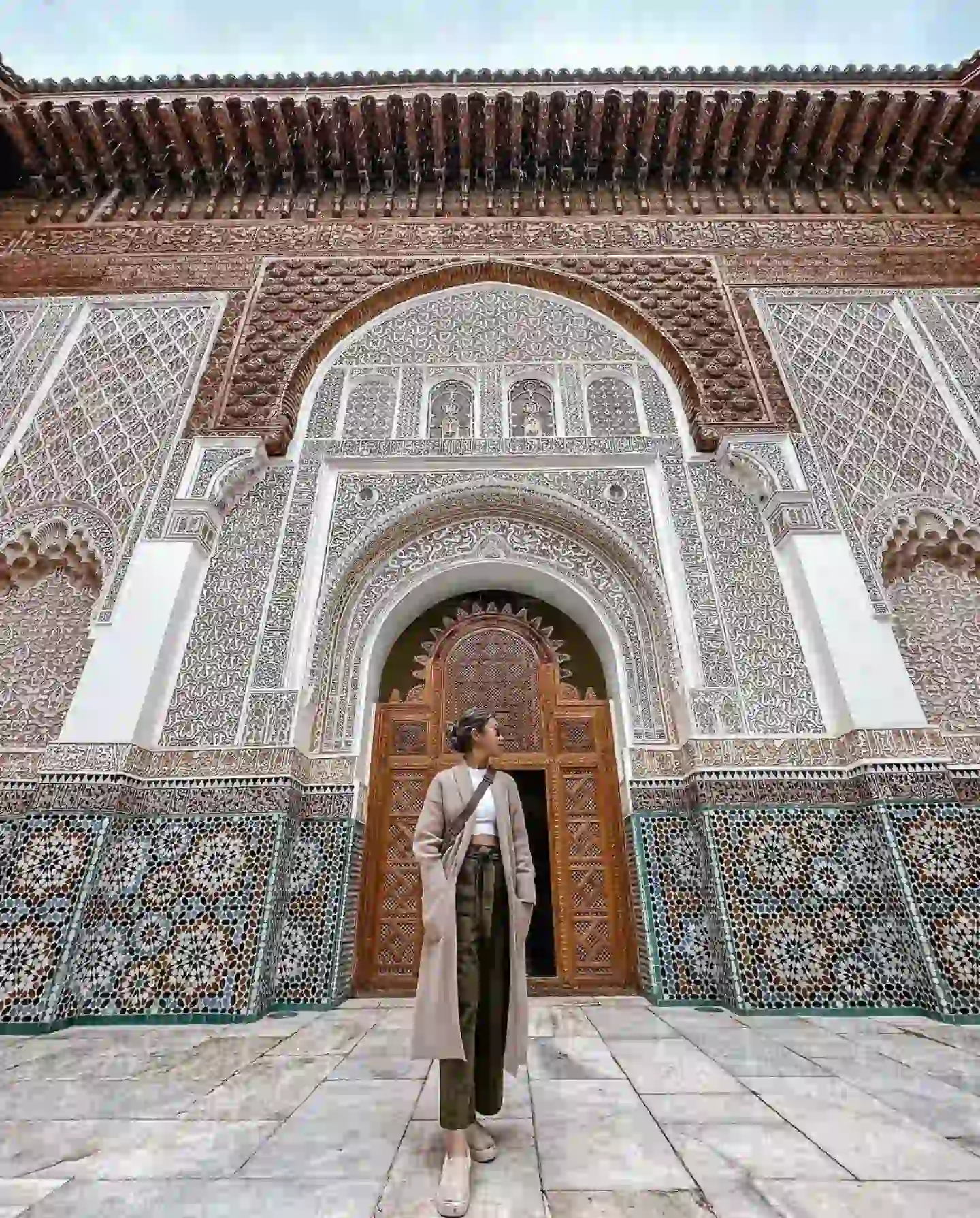 A person in a beige coat and hat stands in front of an intricately designed building with arches and ornate tile work, reminiscent of Marrakesh. The architecture features detailed geometric patterns and traditional motifs. The large wooden doors add a rich, cultural element to the scene.