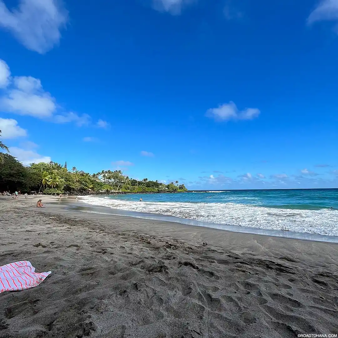 A serene beach in Maui with dark sand, clear blue water, and a slightly cloudy sky. There are a few people in the distance enjoying the beach, and a striped towel is laid near the foreground. Lush greenery, quintessential of Hawaii, is visible in the background.