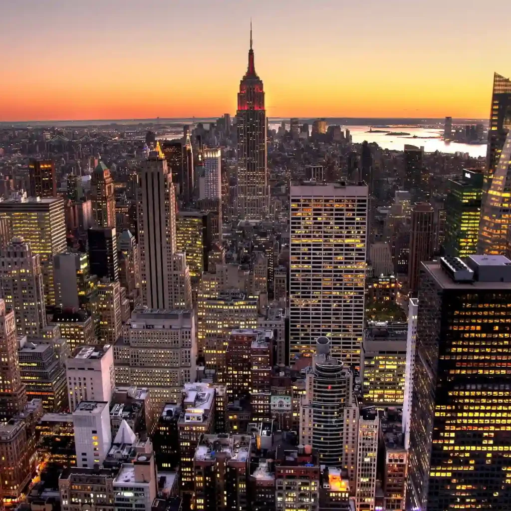 Aerial view of New York City's skyline at dusk, featuring tall skyscrapers with glowing windows. The sky is a gradient of orange to dark blue as the sun sets on the horizon.