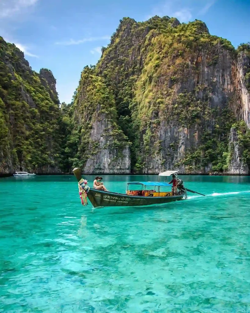 A traditional wooden long-tail boat with passengers moves through clear turquoise waters surrounded by towering limestone cliffs covered in lush greenery, under a bright blue sky. The scene, reminiscent of Phuket's picturesque tropical landscape in Thailand, depicts tranquility at its finest.