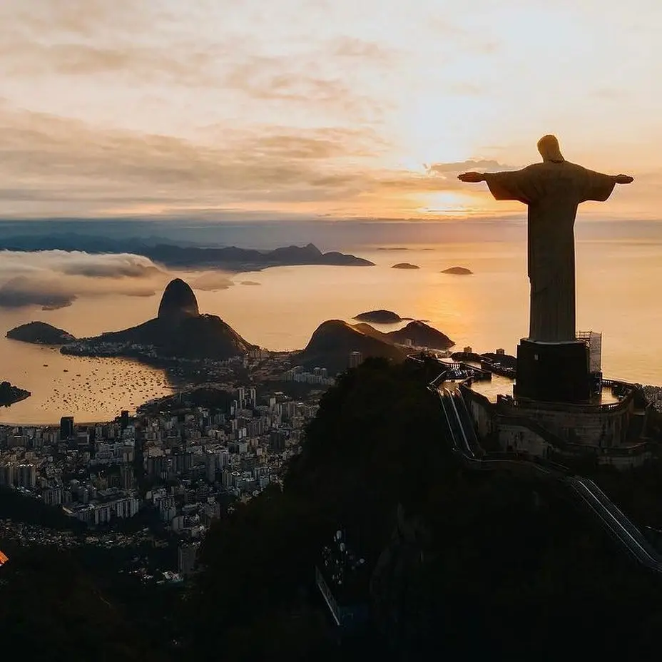 A breathtaking view at sunset of the Christ the Redeemer statue atop Corcovado Mountain in Rio de Janeiro, Brazil. The sprawling cityscape and Sugarloaf Mountain are visible below, with the glowing sun setting over the ocean.