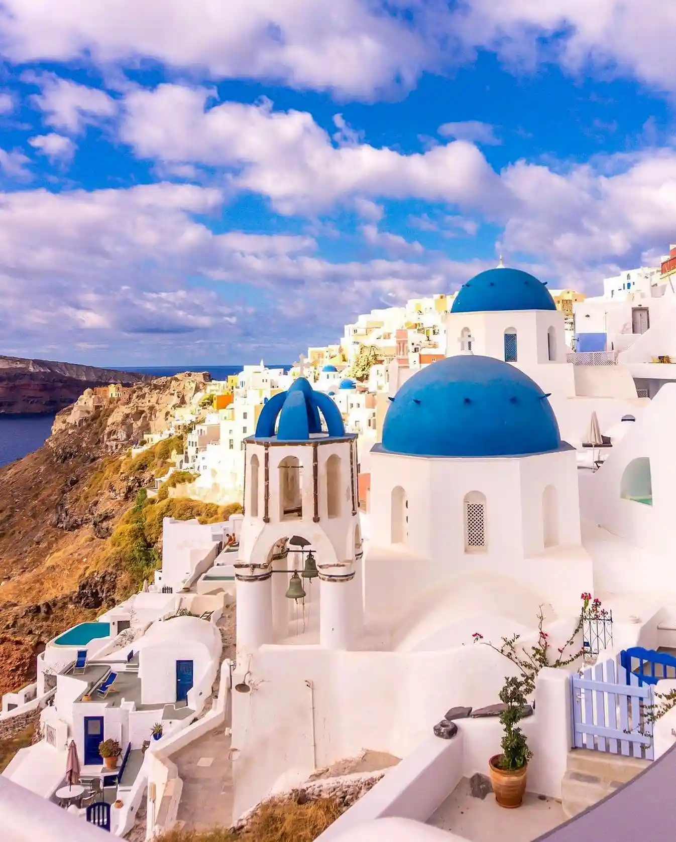 A picturesque view of Santorini, Greece, showcasing its white-washed buildings with iconic blue domes perched on a hillside above the deep blue Aegean Sea. The clear sky is dotted with a few clouds, and vibrant colors adorn the scene, creating a serene atmosphere in Santorini's enchanting landscape.