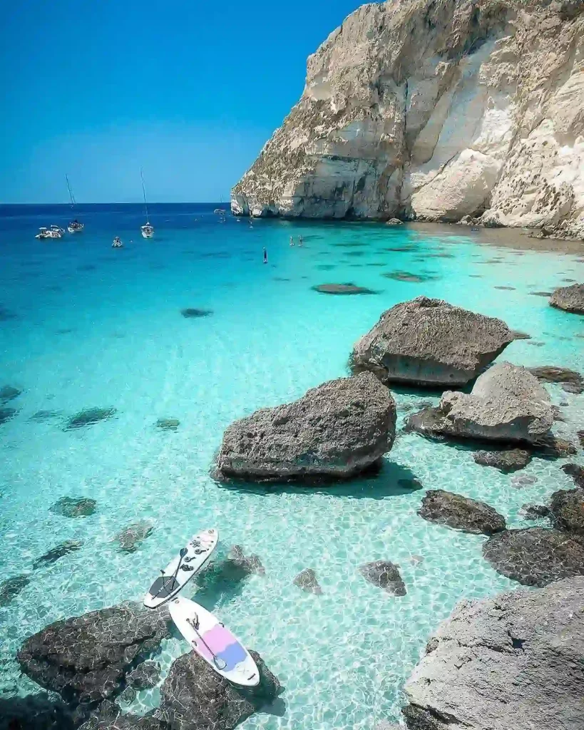 A stunning coastal scene in Sardinia features clear turquoise waters with rocky cliffs in the background. Several boats are anchored in the bay, and two paddleboards rest on the calm, shallow water near large rocks. The sky is clear and bright blue.