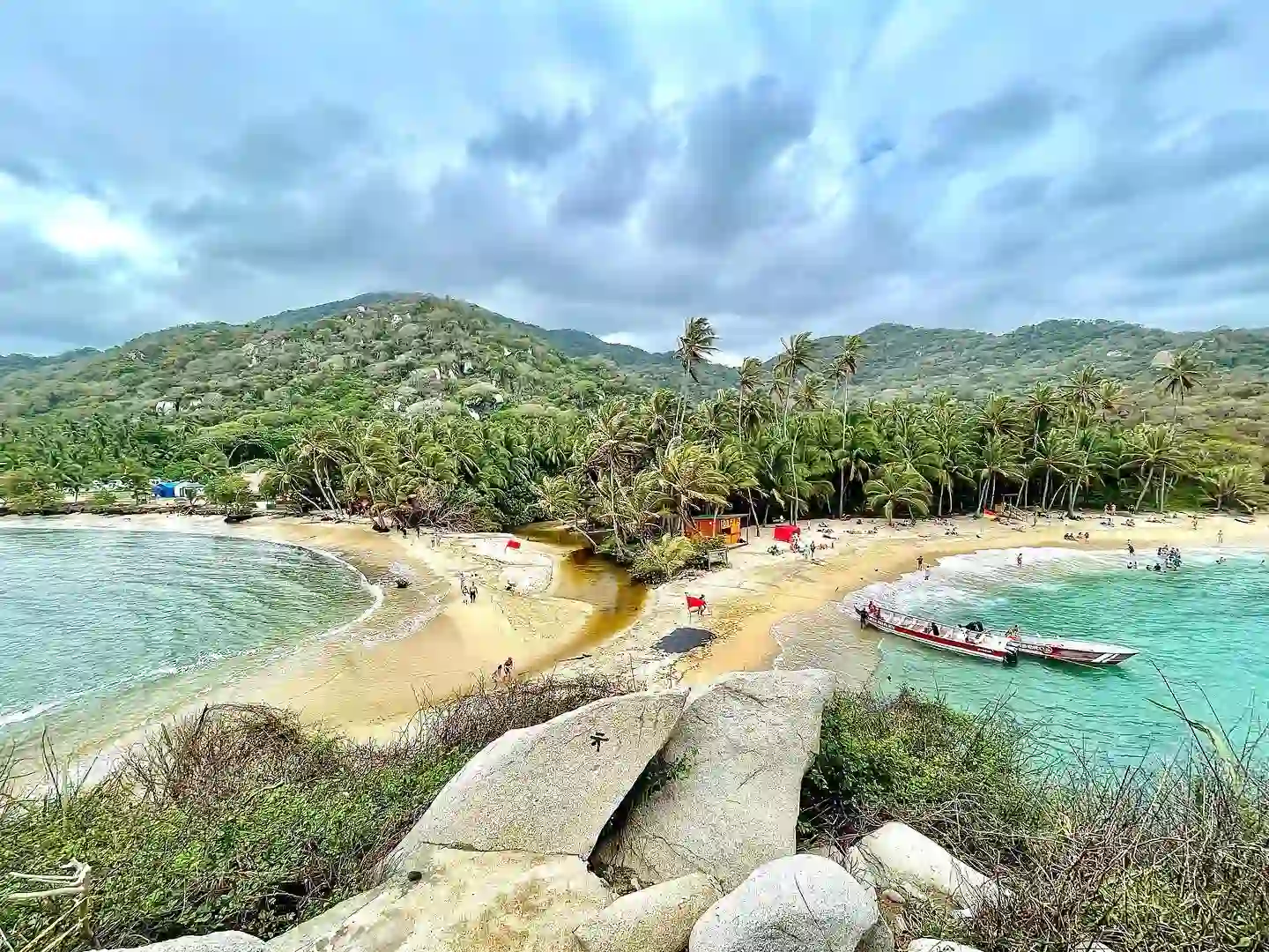 A scenic view of a tropical beach with golden sand and lush green hills in the background. The shoreline curves around a small bay, meeting clear turquoise waters. Boats are docked near the shore, and palm trees line the beach, providing shade to visitors in Tayrona National Park, Colombia.