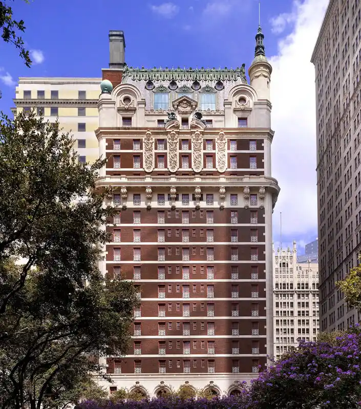 The Adolphus, a multi-story historic building with ornate architecture, stands gracefully between modern structures, with trees in the foreground and a blue sky in the background.