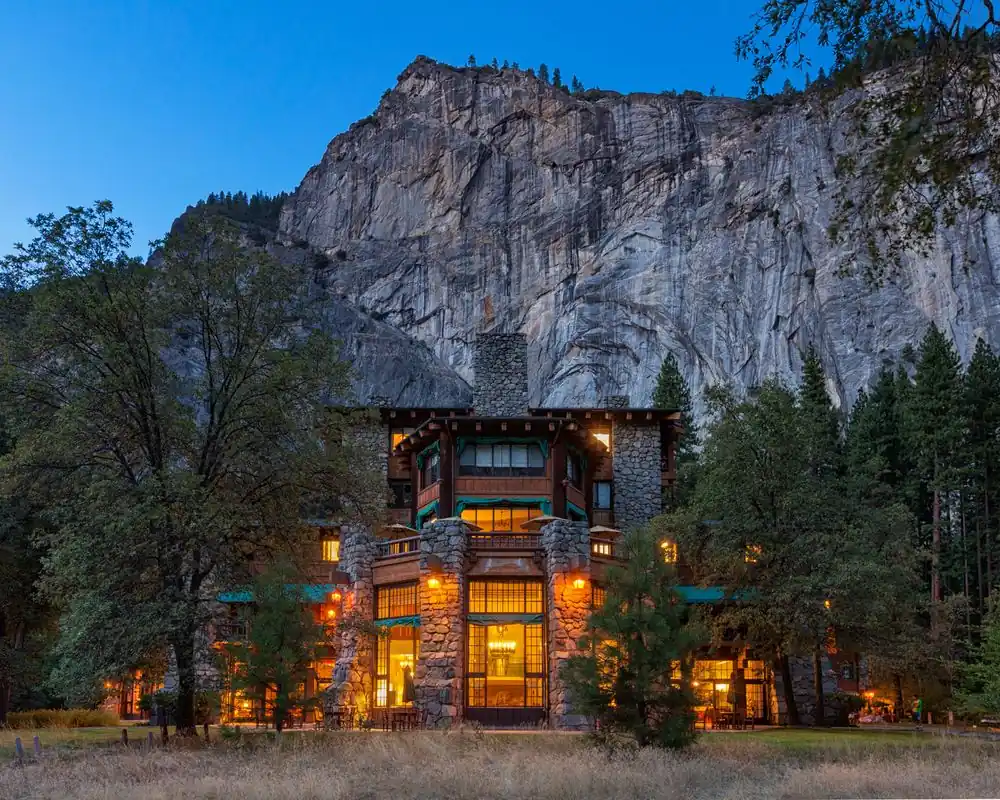 The Ahwahnee, a large multi-story stone building with illuminated windows, is set against a backdrop of a steep rocky mountain and surrounded by trees at dusk.