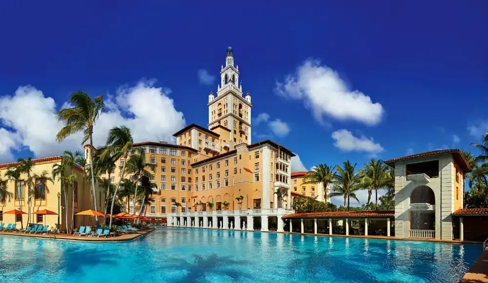 The large, historic Biltmore Hotel with beige and orange tones sits behind a spacious pool under a clear blue sky, flanked by palm trees and lounge chairs.