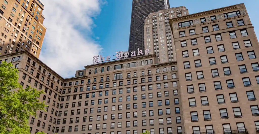 A historic multi-story building in an urban area, adorned with "The Drake" sign on the rooftop, is beautifully framed by taller modern buildings in the background under a partly cloudy sky.