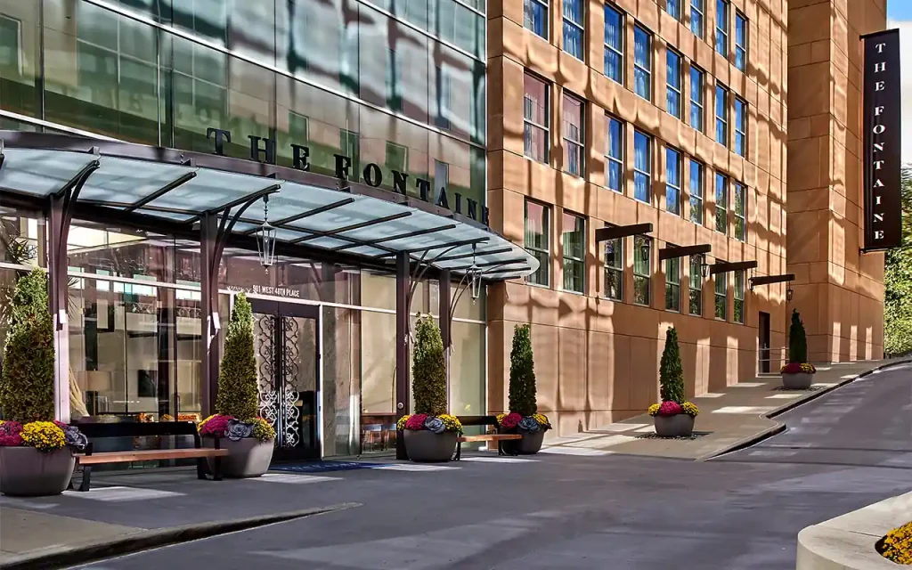 The entrance of The Fontaine, a modern hotel, showcases a sleek glass canopy, elegant potted plants, and a stylish sign on its facade.