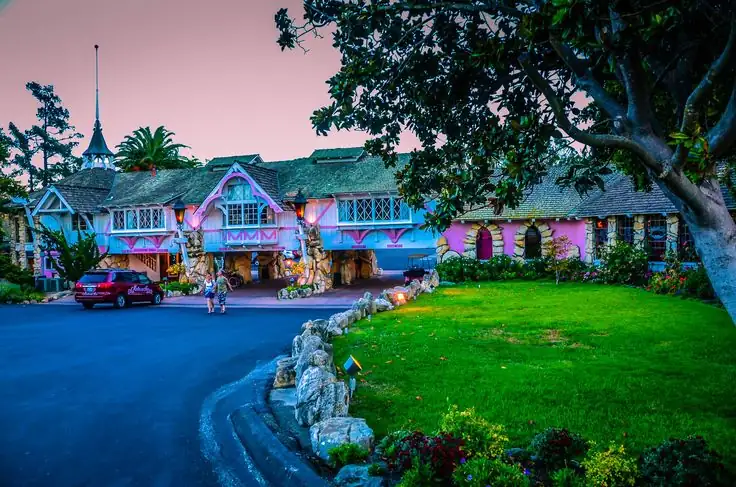 A whimsical building with colorful, fairy-tale-like architecture stands surrounded by greenery and trees. In front, there is a black paved area with a red car and some people walking. The sky above is a soft pink, suggesting early evening or sunrise—reminiscent of The Madonna Inn's charm.