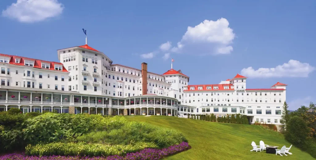 The Omni Mount Washington Resort, a large white hotel with red roofs, stands on a grassy hill with flowerbeds and outdoor seating areas under a clear blue sky.