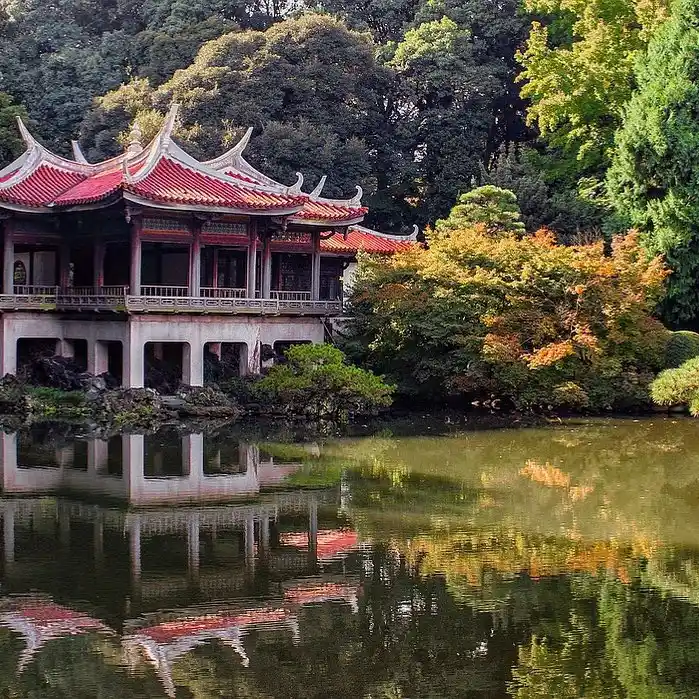 A traditional Japanese building with a red roof is reflected in a serene pond surrounded by lush greenery and autumn-colored trees. The scene is calm and picturesque, capturing the essence of nature and architecture in harmony, reminiscent of a tranquil retreat amidst the bustling life of Tokyo.